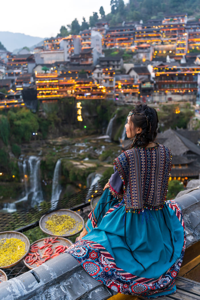 Mujer con traje tradicional frente a un pueblo iluminado en China.